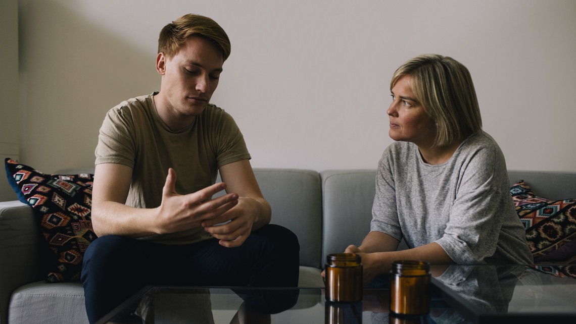 Mature parent looking at sad son sitting on sofa at home