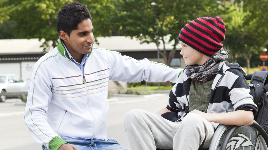 En kille i rullstol och hans assistent som står på knä bredvid. 