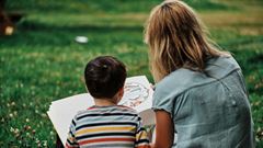 En kvinna sitter utomhus och läser en bok för en pojke.