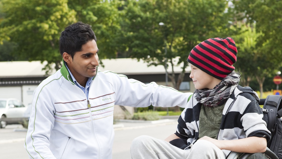 En kille i rullstol och hans assistent som står på knä bredvid. 