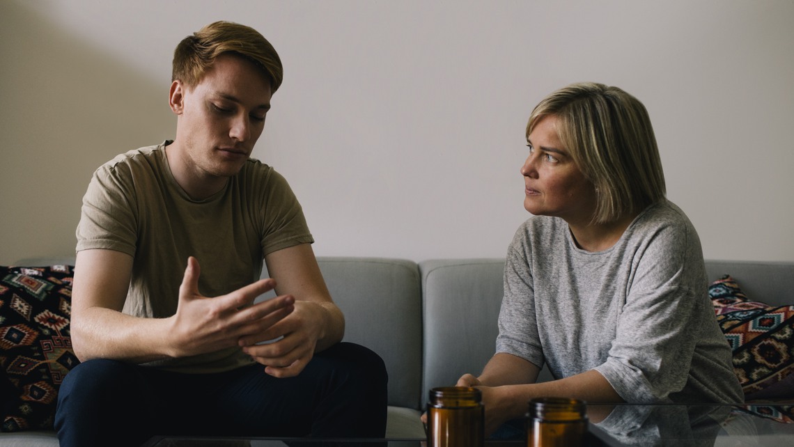 Mature parent looking at sad son sitting on sofa at home