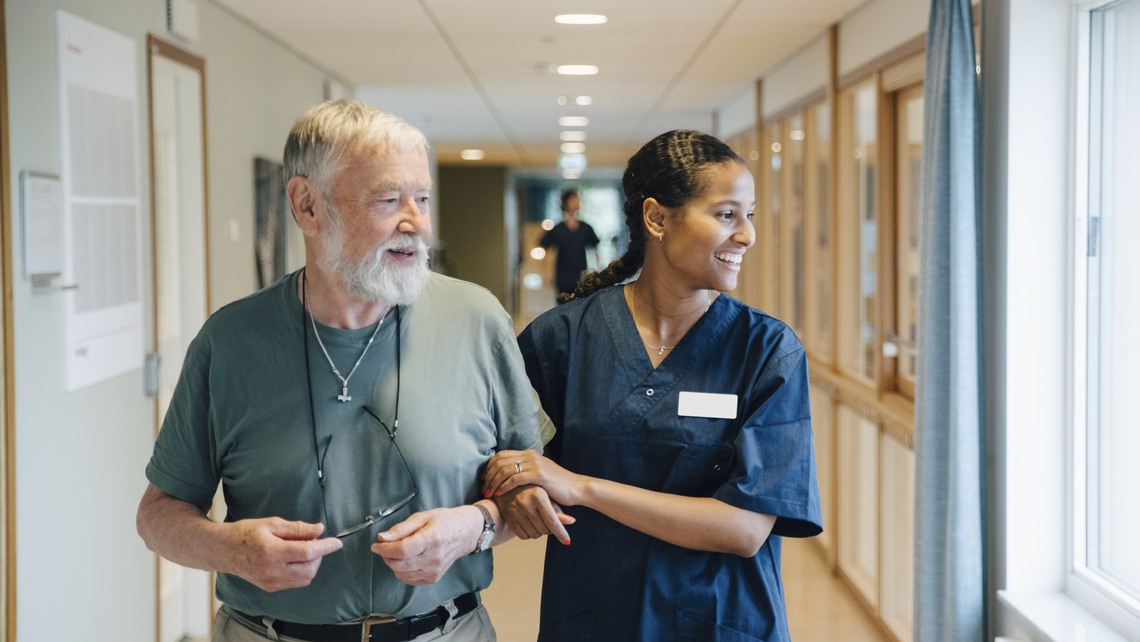 Smiling senior man walking arm in arm with female nurse while looking away in alley at nursing home